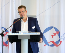 Ein Redner steht am Mikrofonpult vor dem Banner der Veranstaltung und spricht in den Raum.