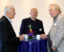 Drei Männer im Gespräch an einem Stehtisch mit Getränken.
