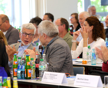 Blick in den Saal mit mehreren Teilnehmenden an Konferenztischen im Gespräch.