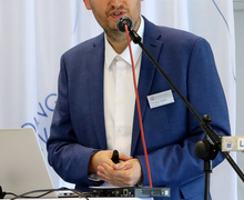 Redner am Laptop spricht am Pult vor dem Hintergrund eines Veranstaltungslogos.