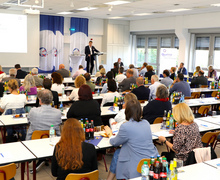 Ein Redner steht vorne im Saal am Pult, das Publikum verfolgt seinen Vortrag.