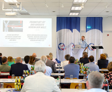 Ein Redner steht links vor einer großen Leinwand mit projizierter Präsentation.