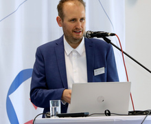 Redner am Laptop spricht am Pult vor dem Hintergrund eines Veranstaltungslogos.